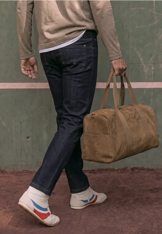 Person in Jeans und Sneakern hält eine braune Reisetasche vor grüner Wand.