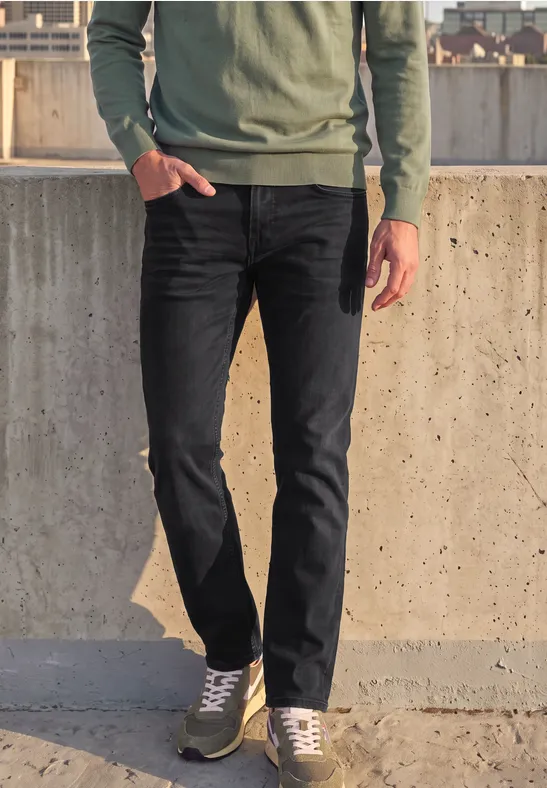 Eine Person in schwarzer Jeans, grauen Sneakern und grünem Pullover lehnt an einer Betonmauer im Freien.