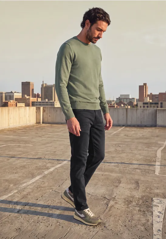 Ein Mann in grünem Pullover und dunkler Hose geht auf einem Parkdeckdach mit Stadtkulisse.