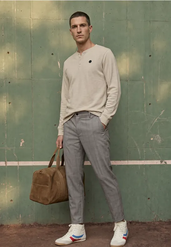 Ein Mann in beigem Pullover, grauen Hosen und Sneakers steht vor grüner Wand und trägt eine braune Reisetasche.