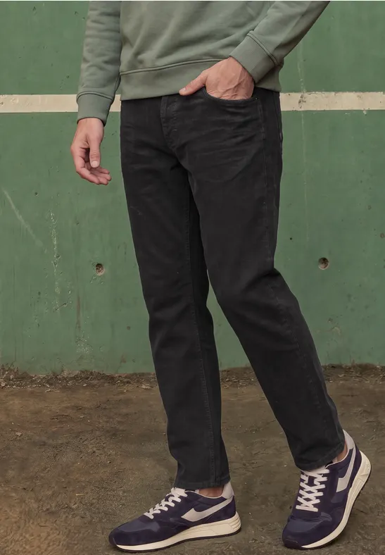 Eine Person trägt ein grünes Oberteil, schwarze Jeans und blaue Sneaker vor einer grünen Wand.