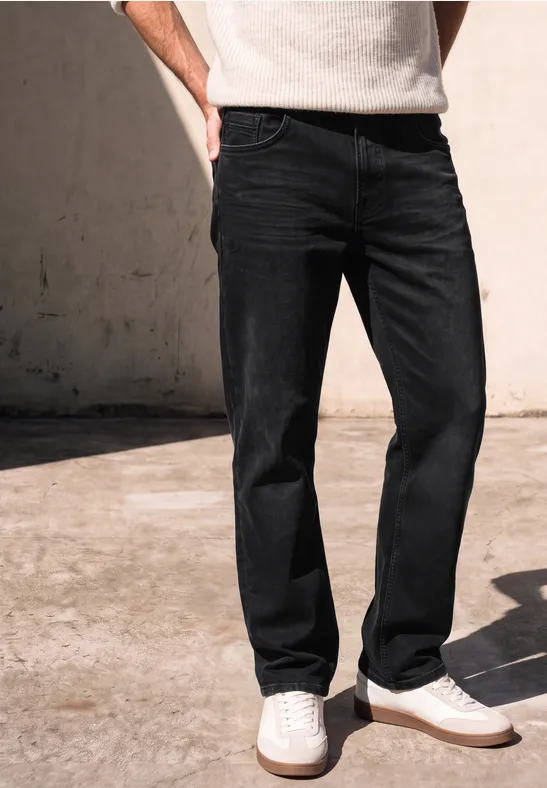 Person steht vor Betonwand, trägt schwarze Jeans und weiße Sneaker.