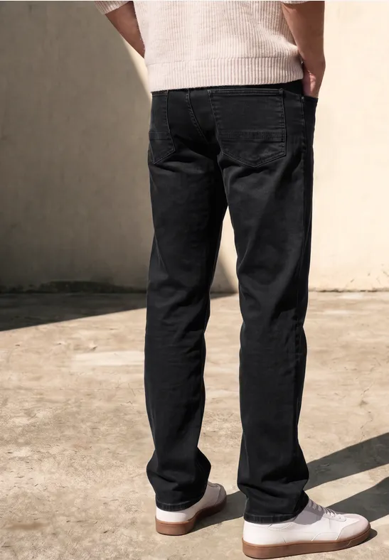Rückansicht einer Person in schwarzen Jeans und weißen Slip-on-Schuhen auf hellem Betonboden.