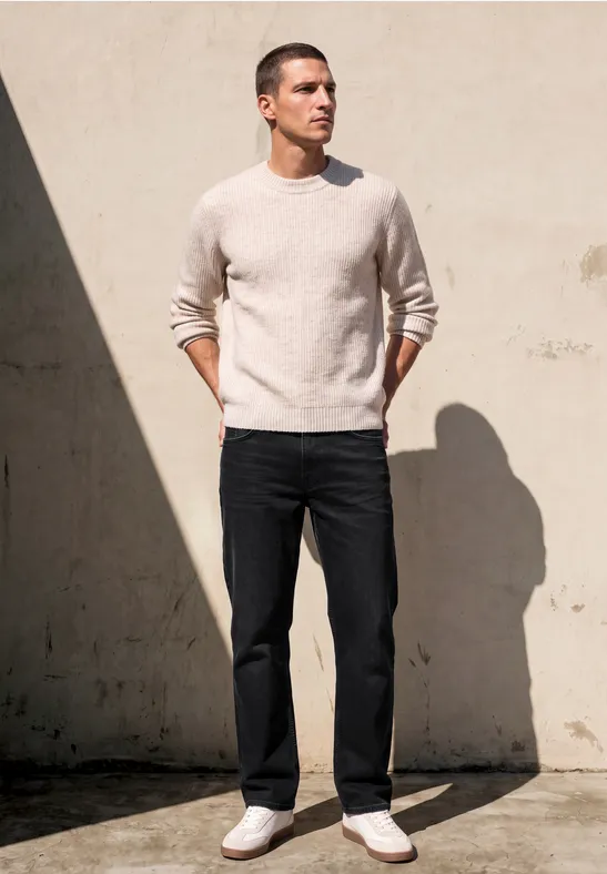 Ein Mann steht vor einer hellen Wand im Schatten, trägt beigen Pullover, dunkle Jeans und weiße Sneakers.