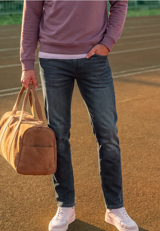 Person in Jeans, weißen Sneakern und rosa Sweatshirt hält braune Reisetasche auf Laufbahn.