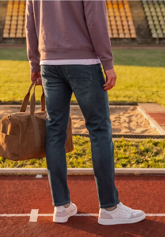 Person in Jeans, weißen Sneakern und Pullover trägt Reisetasche am Sportplatzrand.