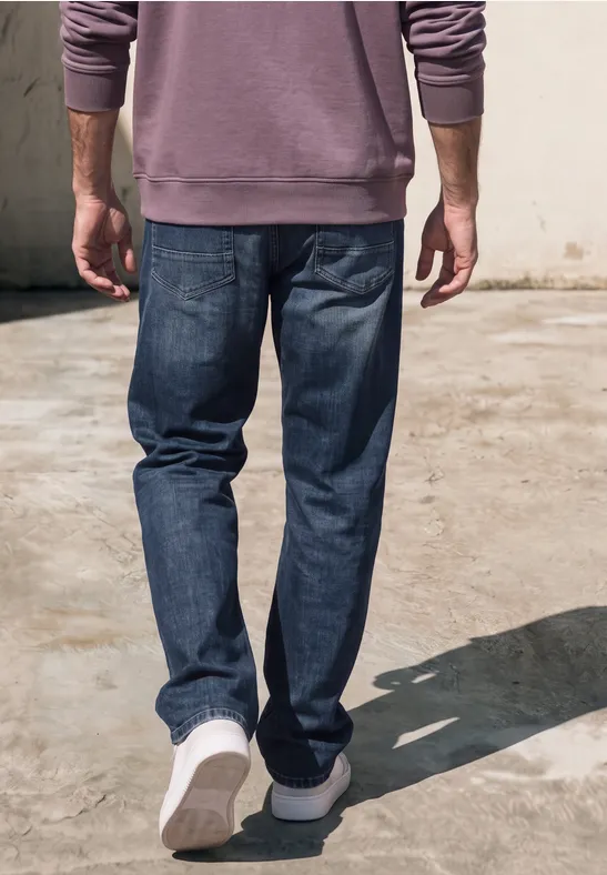Ein Mann spaziert weg, trägt blaue Jeans, weiße Sneaker und einen lila Pullover.