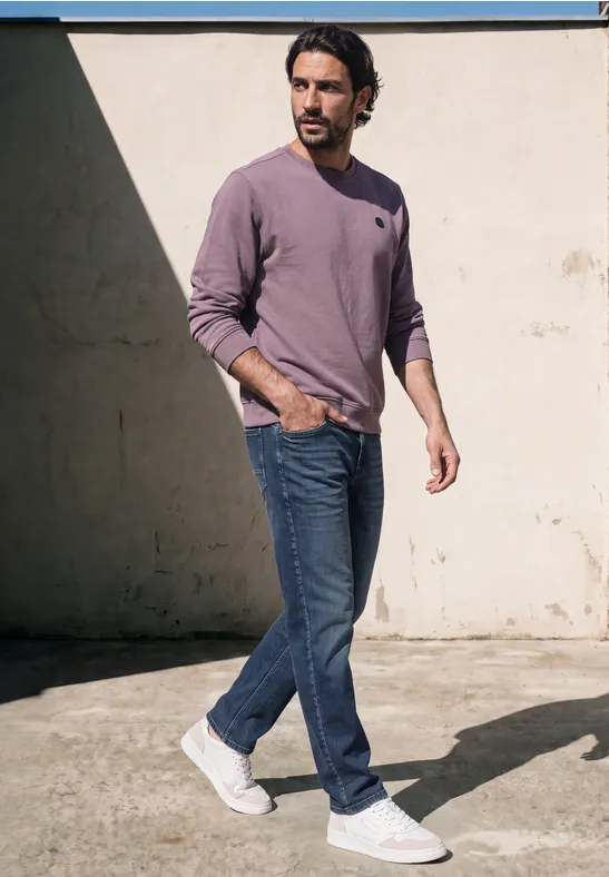 Ein Mann in lila Pullover, Jeans und weißen Sneakern geht vor einer hellen Wand spazierend.