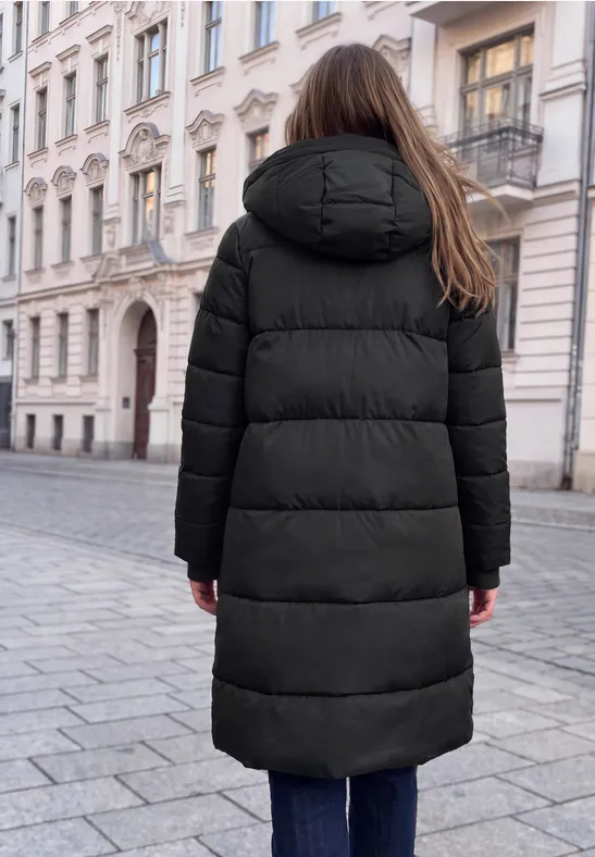 Frau in schwarzer Steppjacke von hinten auf Kopfsteinpflaster vor historischen Häusern.