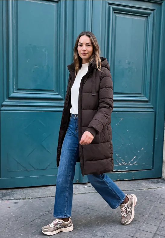 Junge Frau in Jeans, weißem Shirt und langer brauner Daunenjacke spaziert vor einer türkisfarbenen Tür.