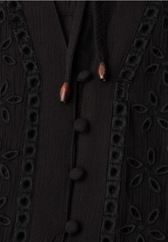 Nahaufnahme eines schwarzen Kleidungsstücks mit Stoffknöpfen, Holz-Kordeln und filigranen Lochstickereien.