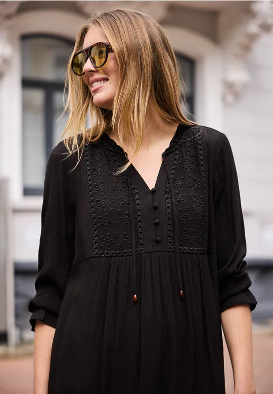 Frau mit schwarzem, besticktem Maxikleid und langen Ärmeln steht vor einem Gebäude, Gesicht unscharf.