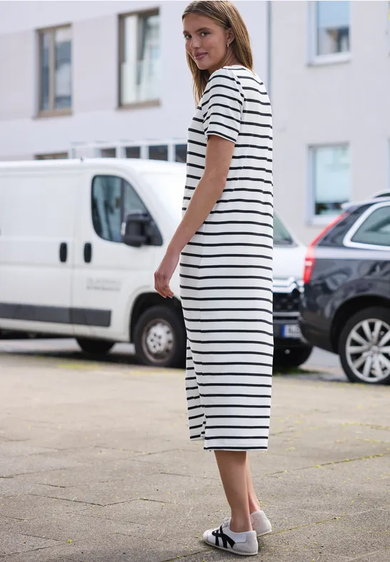Vrouw in witte, zwart gestreepte midijurk en witte sandalen staat op stoep voor geparkeerde auto’s.