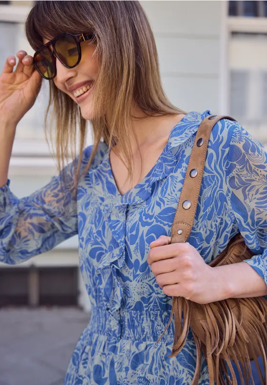 Vrouw draagt blauw, gemêleerd topje met taille-inzet en schoudertas, staat voor gevel.