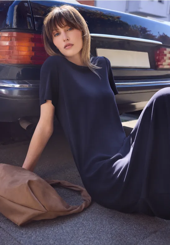 Een vrouw zit op de grond naast een geparkeerde auto, draagt een lange donkere jurk en heeft een tas naast zich.