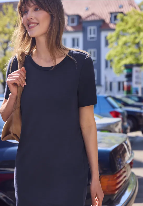 Vrouw in zwart T-shirtjurkje met schoudertas, lopend door een zonnige stadsstraat.