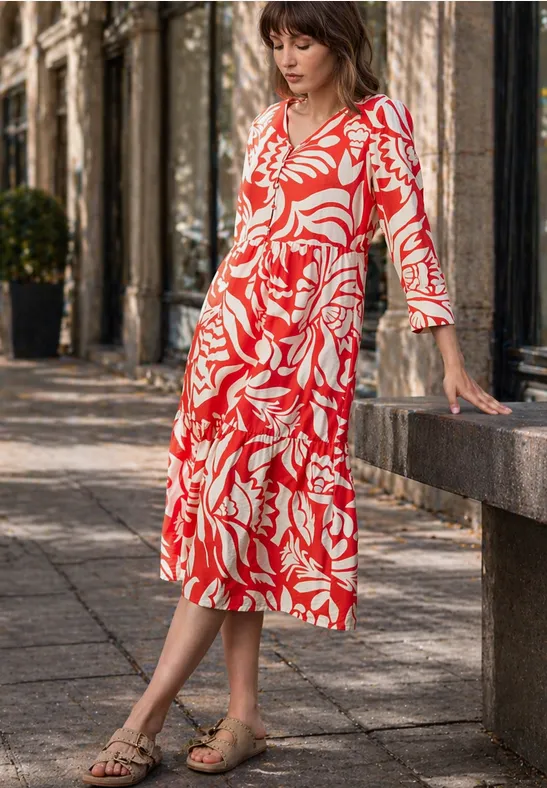 Eine Frau in rotem weißem Blumenmuster-Midikleid steht draußen an einer Steinbank.