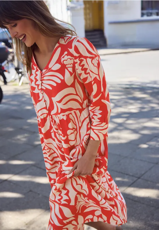Vrouw in een rood-wit bloemenjurk, lopend op straat op een zonnige dag.