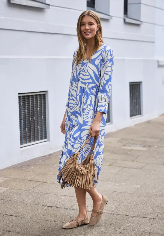 Frau in blau-weiß floralem Midikleid, mit Basttasche und Espadrilles vor hellem Haus