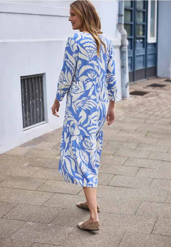 Eine Frau spaziert auf dem Bürgersteig in einem blau-weißen, floralen Maxikleid und braunen Slippern.