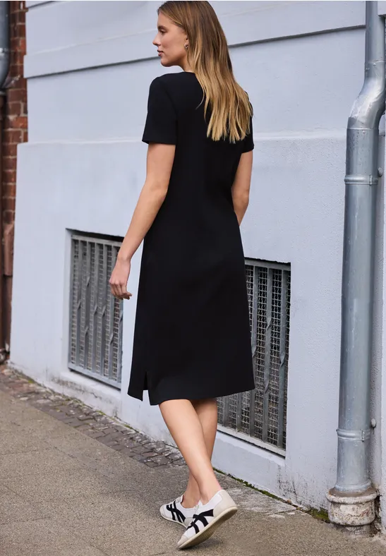 Vrouw in zwarte knie-lange jurk en sneakers loopt langs een grijze gevel met een ventilatierooster.