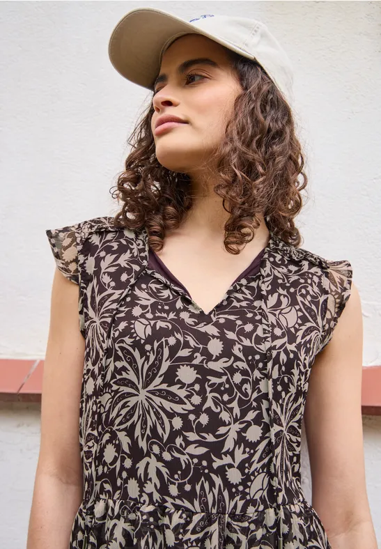Frau in ärmellosem, braunem Kleid mit floralem Muster vor heller Wand.