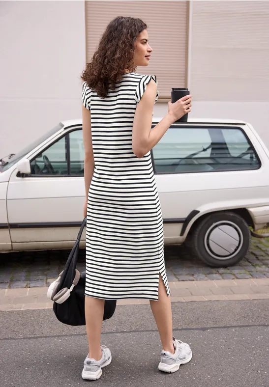 Frau im gestreiften Kleid mit Kaffee in der Hand vor einem weißen Kombi auf dem Gehweg.