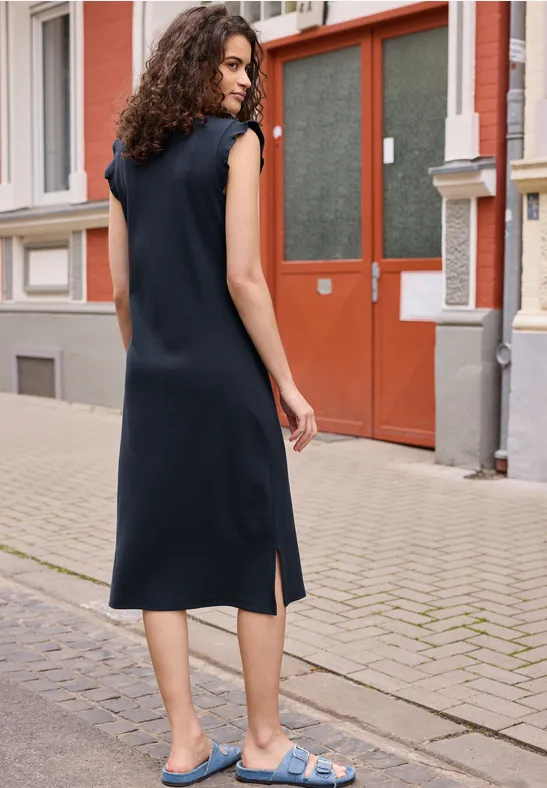 Een vrouw in een mouwloze zwarte jurk en blauwe sandalen staat voor een rode deur.