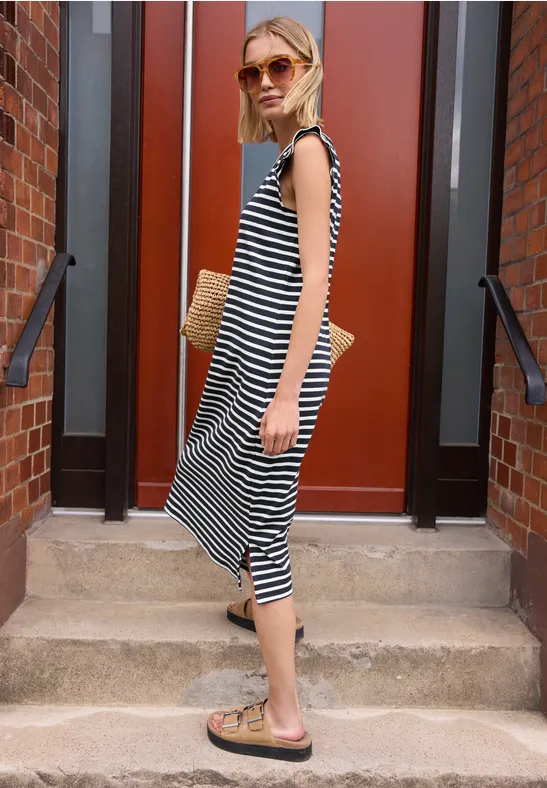 Frau in gestreiftem Midi-Kleid und Sandalen hält einen Strohtasche auf einer Treppe.