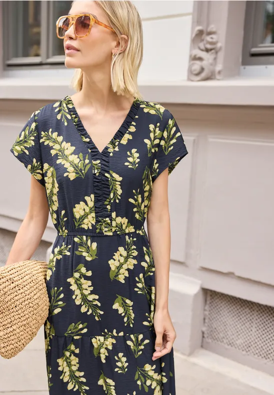Frau trägt ein schwarzes, florales Wickelkleid mit grünen Blättern und hält einen Strohkorb.