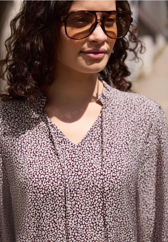 Frau mit lockigem Haar trägt eine helle Bluse mit kleinem Blumenmuster und V-Ausschnitt.