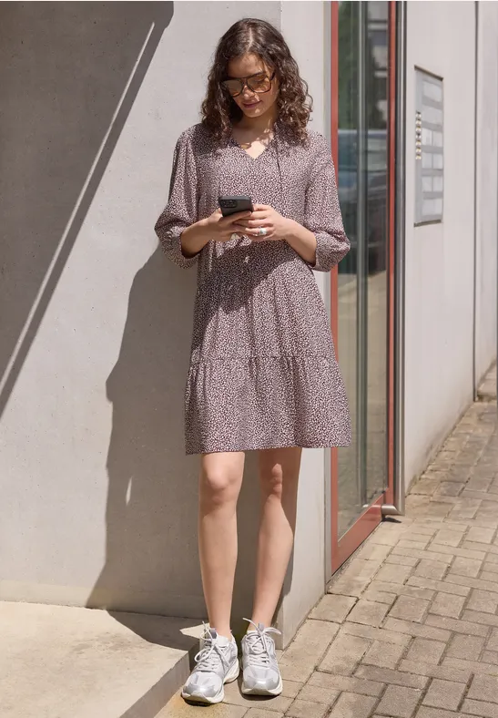 Frau in lila gepunktetem Kleid und weißen Sneakern steht draußen an einer Hauswand und hält ein Handy.