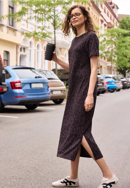 Eine Frau in schwarzem gemustertem Midikleid und weißen Sandalen hält einen Coffee-to-go-Becher auf der Straße.