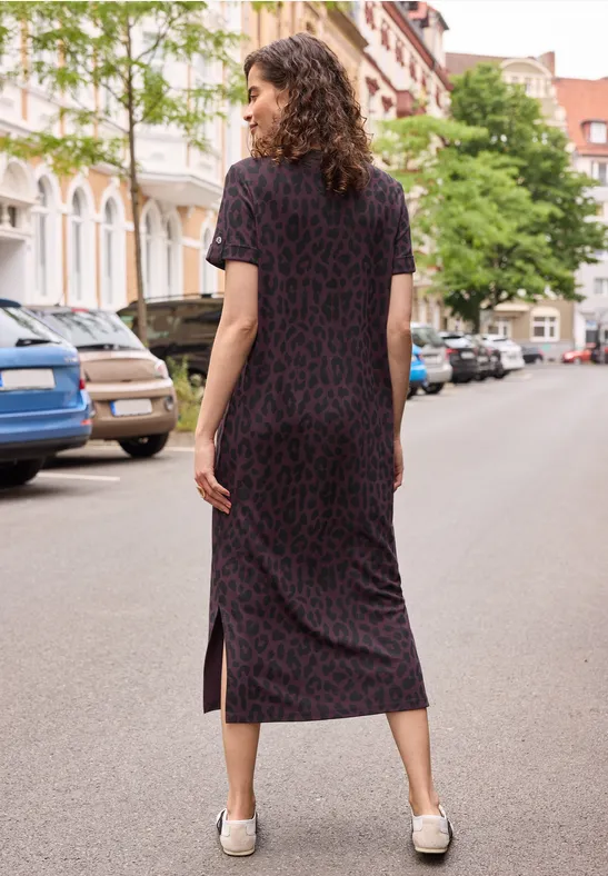 Frau in gemustertem schwarzen Midi-Kleid und weißen Loafern steht auf ruhiger Stadtstraße, von hinten.