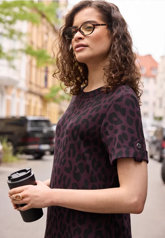 Junge Frau mit lockigen Haaren hält einen Coffee-to-go-Becher, trägt lässiges Leoparden-Print-Shirt in der Stadt.