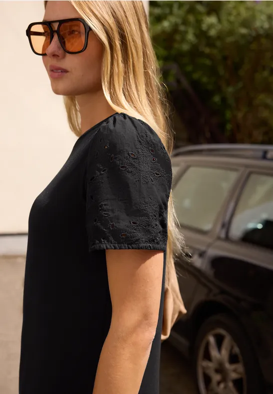 Eine Frau mit langen blonden Haaren in schwarzem Kleid steht seitlich vor einem geparkten schwarzen Auto.
