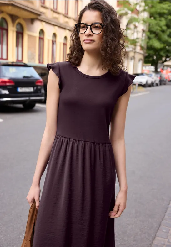 Frau in ärmellosem schwarzen Kleid steht auf Gehweg, im Hintergrund Häuser und parkende Autos.