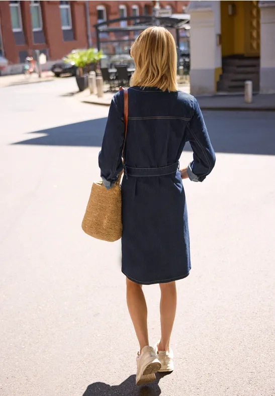 Frau in jeansblauem Kleid geht mit Strohtasche über eine sonnige Straße.