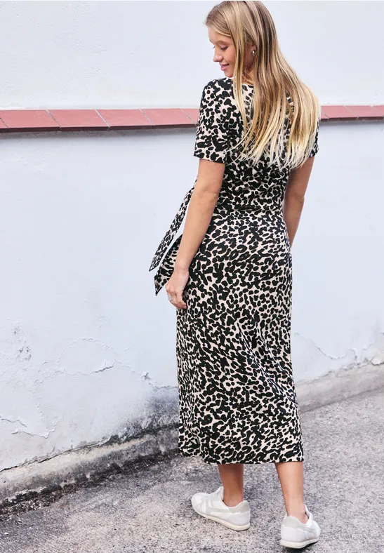Eine blonde Frau in einem gemusterten Midikleid und weißen Sneakern geht an einer hellen Wand entlang.