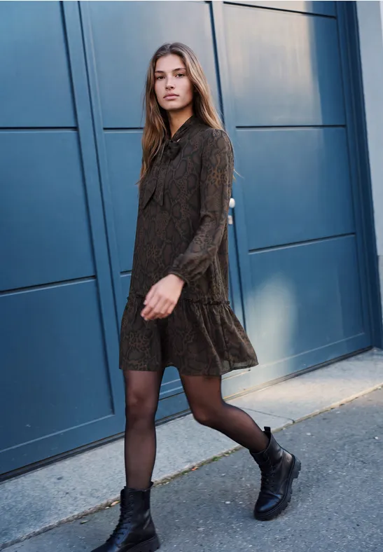 Eine Frau in schwarzem Minikleid, Strumpfhose und Boots spaziert vor einer blauen Garagentür.