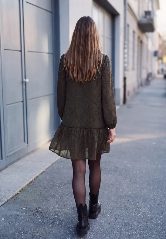Frau mit braunem Haar, dunklem Kleid und Stiefeln geht eine ruhige Stadtstraße entlang.