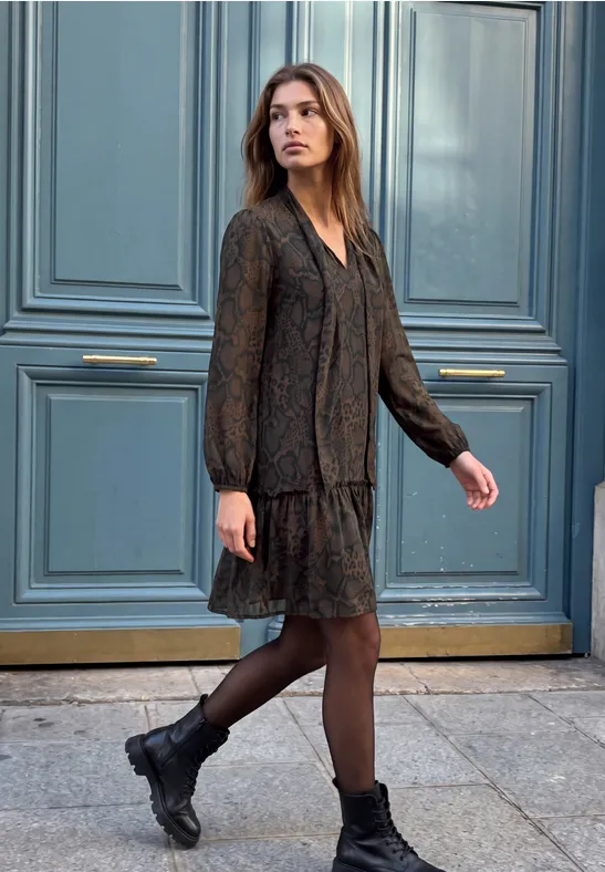 Junge Frau in schwarzem, langärmligem Kleid und Boots steht vor einer blauen Tür auf dem Bürgersteig.