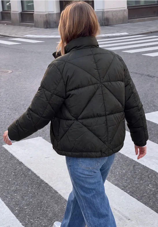 Eine Person von hinten in grüner Steppjacke und blauen Jeans beim Überqueren eines Zebrastreifens.