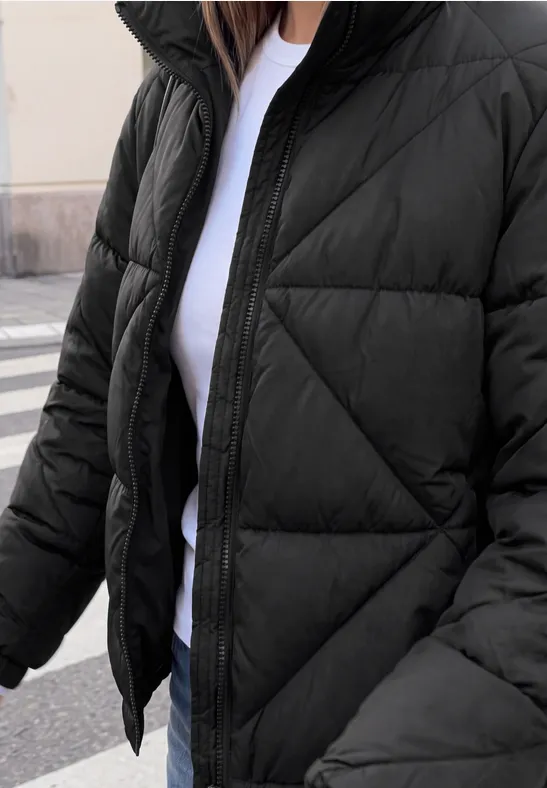 Person trägt schwarzen Steppmantel über weißem T-Shirt, dazu Jeans, steht auf Gehweg.