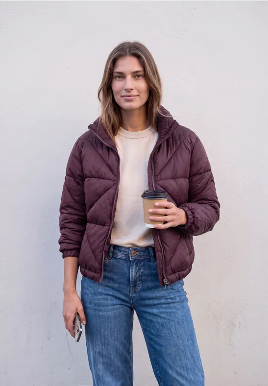 Jonge vrouw voor een witte muur, glimlacht, draagt paarse gewatteerde jas en jeans, met koffie en telefoon.