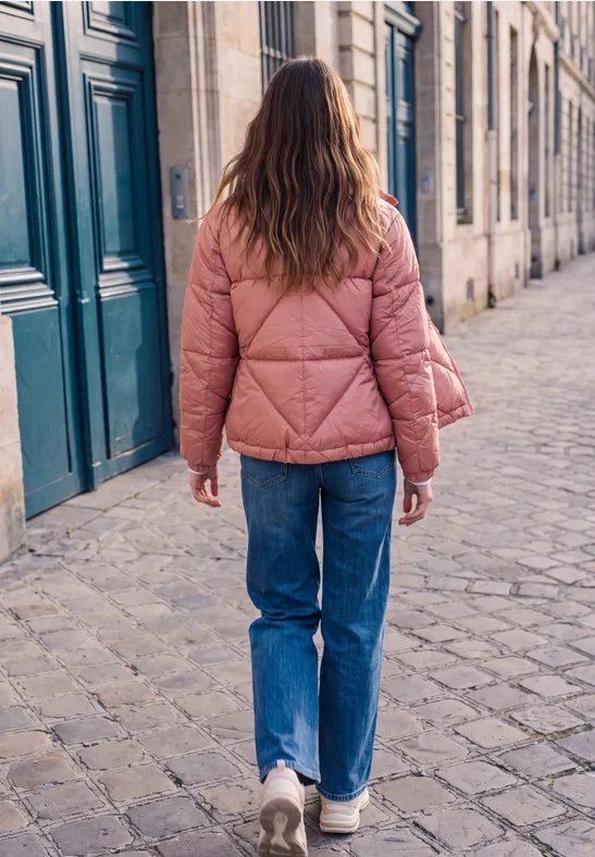 Eine Person geht mit rosa Steppjacke, Jeans und weißen Sneakers auf einer Kopfsteinpflasterstraße spazieren.