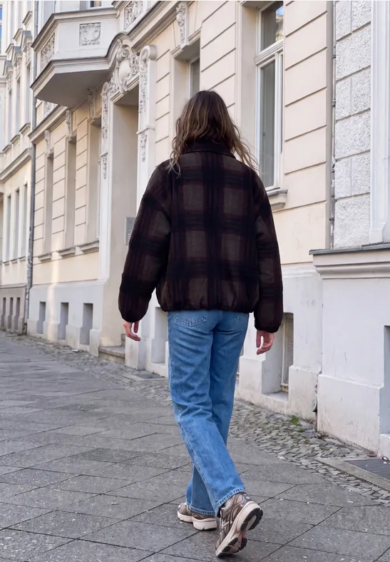 Eine Person in kariertem Mantel, Jeans und Turnschuhen läuft einen Gehweg entlang, vor historischen Häusern.