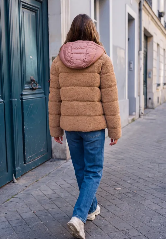 Eine Person in brauner Steppjacke mit rosa Kapuze, blauen Jeans und Sneakers läuft eine gepflasterte Straße entlang.