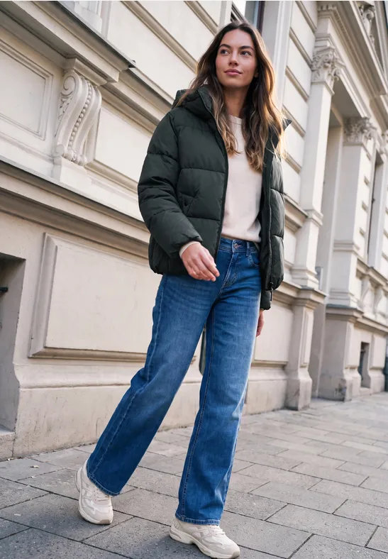 Junge Frau in schwarzer Steppjacke, Jeans und weißen Sneakern läuft an einem klassisch verzierten Haus vorbei.
