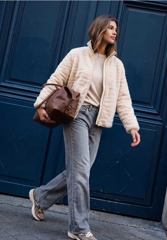 Une femme en jean, veste crème et sac à bandoulière marche devant une porte bleu foncé.
